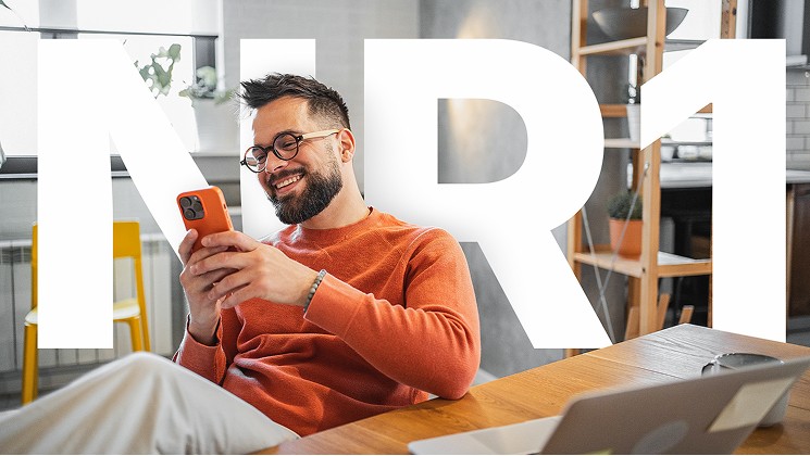 Homem sorrindo ao usar o celular em casa, sobreposto pela sigla NR-1